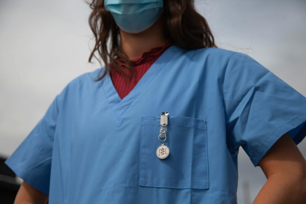 A person wearing blue medical scrubs and a face mask stands outdoors.